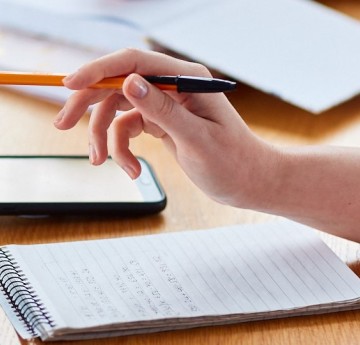 A hand holding a pen hovering over A5 pad of lined paper