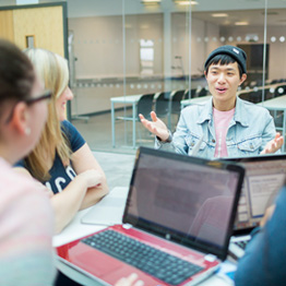 Smiling students sitting around a table with an open laptop on it talking