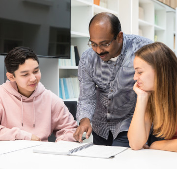 A lecturer going through a notebook with two students who are sitting and listening