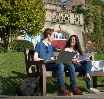 One male on a laptop and one female with a booklet, sitting on a bench outside the Abbey on Singleton Campus surrounded by trees. 